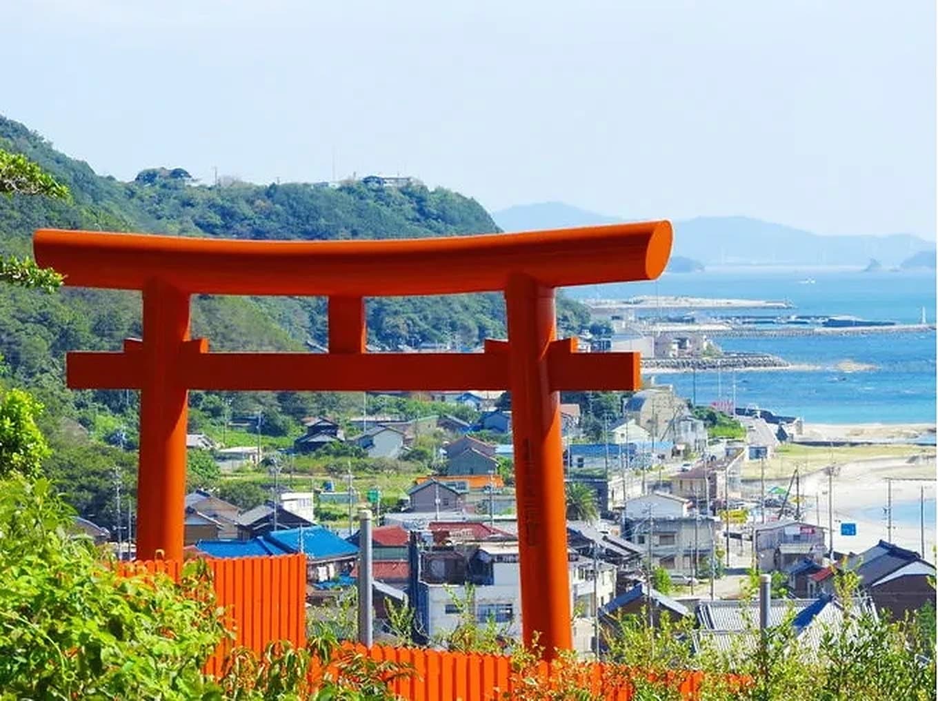A vermilion torii gate above a seaside town, green hills and the blue sea beyond